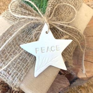 A gift wrapped in brown paper and burlap, tied with twine, and decorated with a sprig of green foliage. A white star-shaped tag with the word "PEACE" is attached. The gift is presented on a wooden surface.