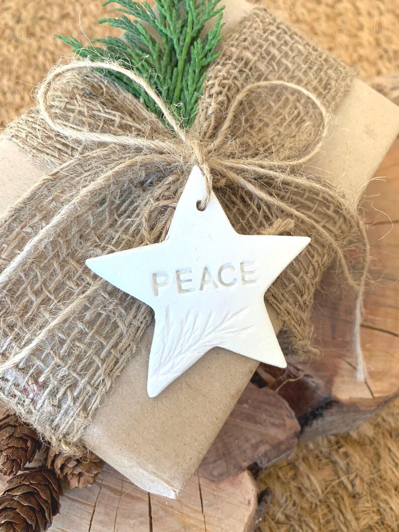 A gift wrapped in brown paper and burlap, tied with twine, and decorated with a sprig of green foliage. A white star-shaped tag with the word "PEACE" is attached. The gift is presented on a wooden surface.