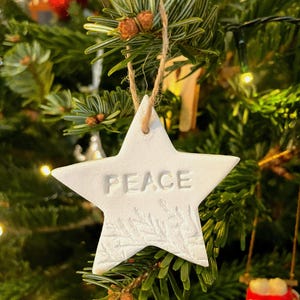 A white star-shaped Christmas decoration with the word "PEACE" embossed on it. The ornament features a textured surface and a leaf design. It hangs from a natural twine string against a Christmas tree background.
