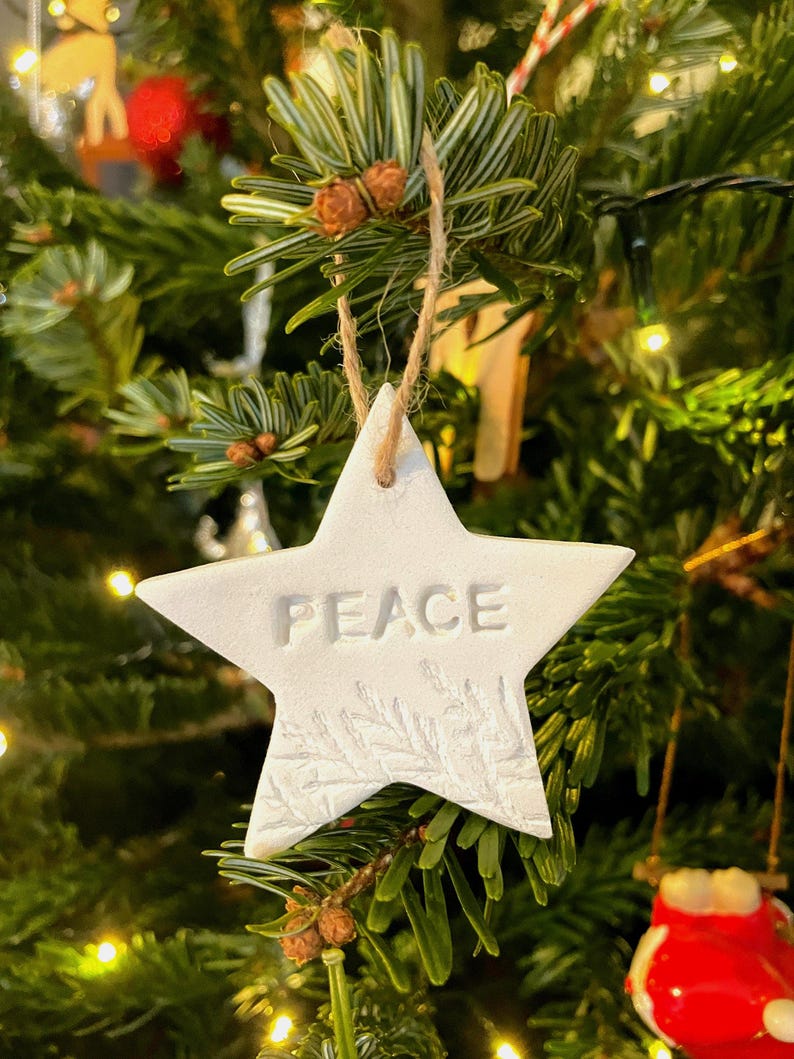 A white star-shaped Christmas decoration with the word "PEACE" embossed on it. The ornament features a textured surface and a leaf design. It hangs from a natural twine string against a Christmas tree background.
