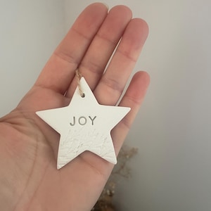 A white star-shaped ornament with the word "JOY" embossed in the centre. The ornament has a textured surface and a small hole at the top for hanging with natural twine. The ornament is held in a hand.