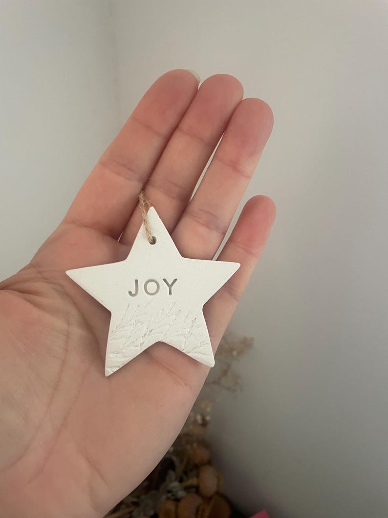 A white star-shaped ornament with the word "JOY" embossed in the centre. The ornament has a textured surface and a small hole at the top for hanging with natural twine. The ornament is held in a hand.