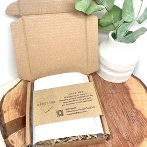 An open cardboard box containing a wrapped item, set on a wooden surface. A small card with text, a heart graphic, and social media details is visible. A white vase with green leaves is in the background.