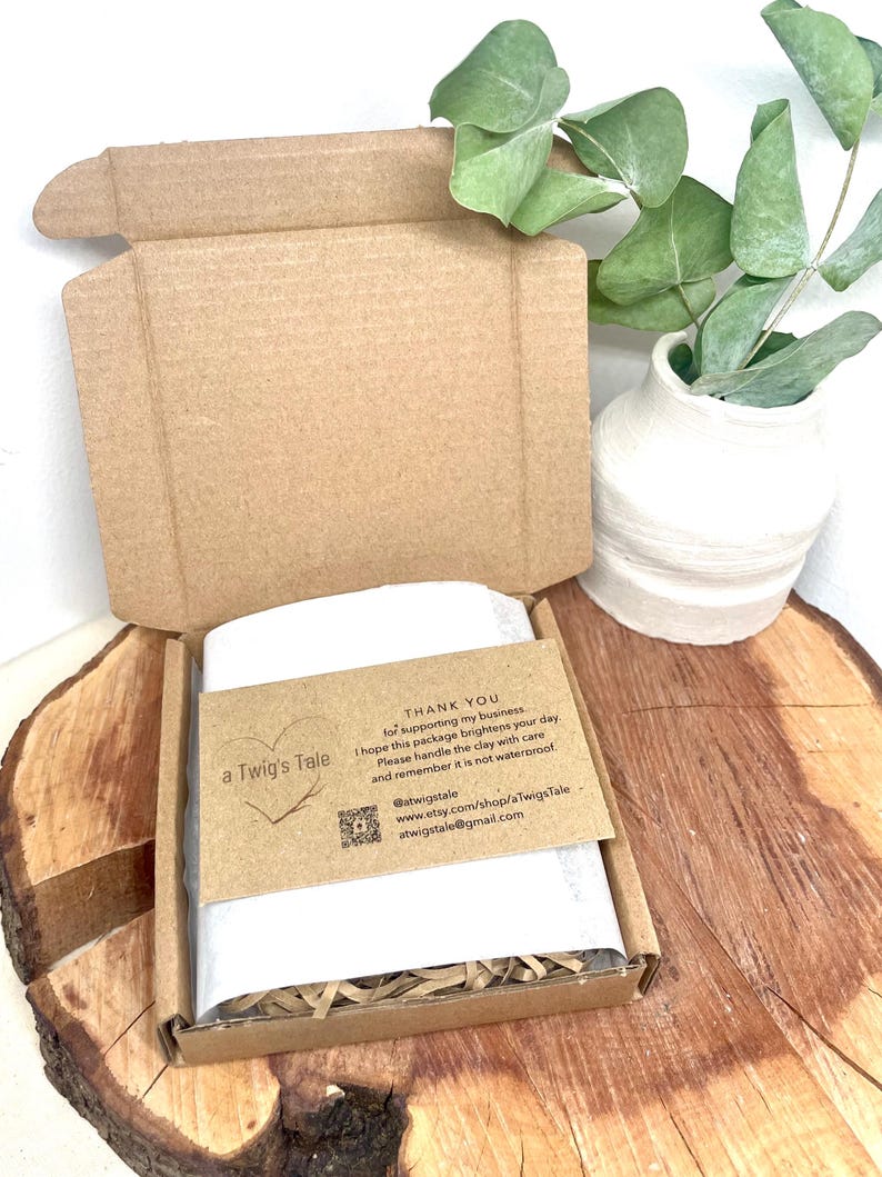 An open cardboard box containing a wrapped item, set on a wooden surface. A small card with text, a heart graphic, and social media details is visible. A white vase with green leaves is in the background.