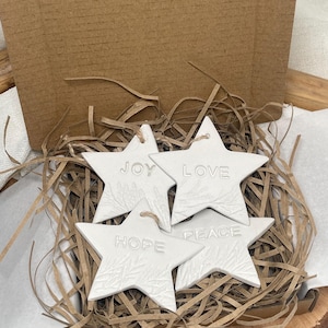 Four white star-shaped ornaments with the words "JOY", "LOVE", "HOPE", and "PEACE" embossed. Each star has a textured sprig design and a twine loop. They are in brown paper filler inside a cardboard box.