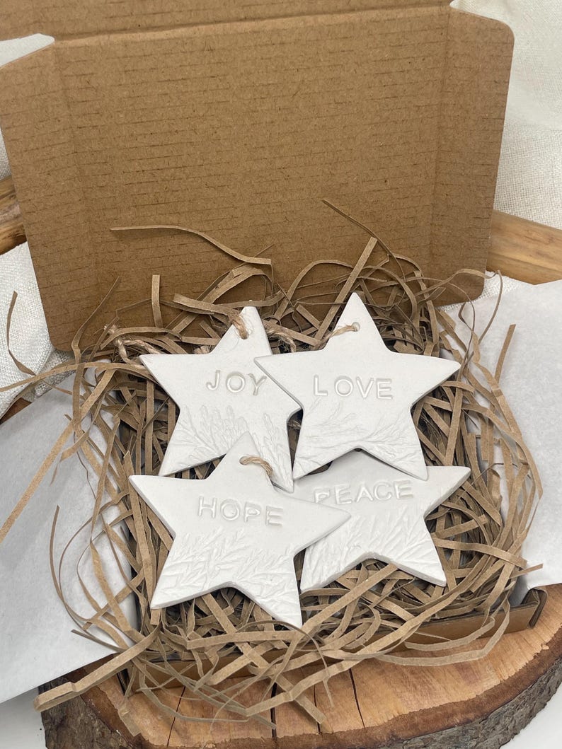 Four white star-shaped ornaments with the words "JOY", "LOVE", "HOPE", and "PEACE" embossed. Each star has a textured sprig design and a twine loop. They are in brown paper filler inside a cardboard box.