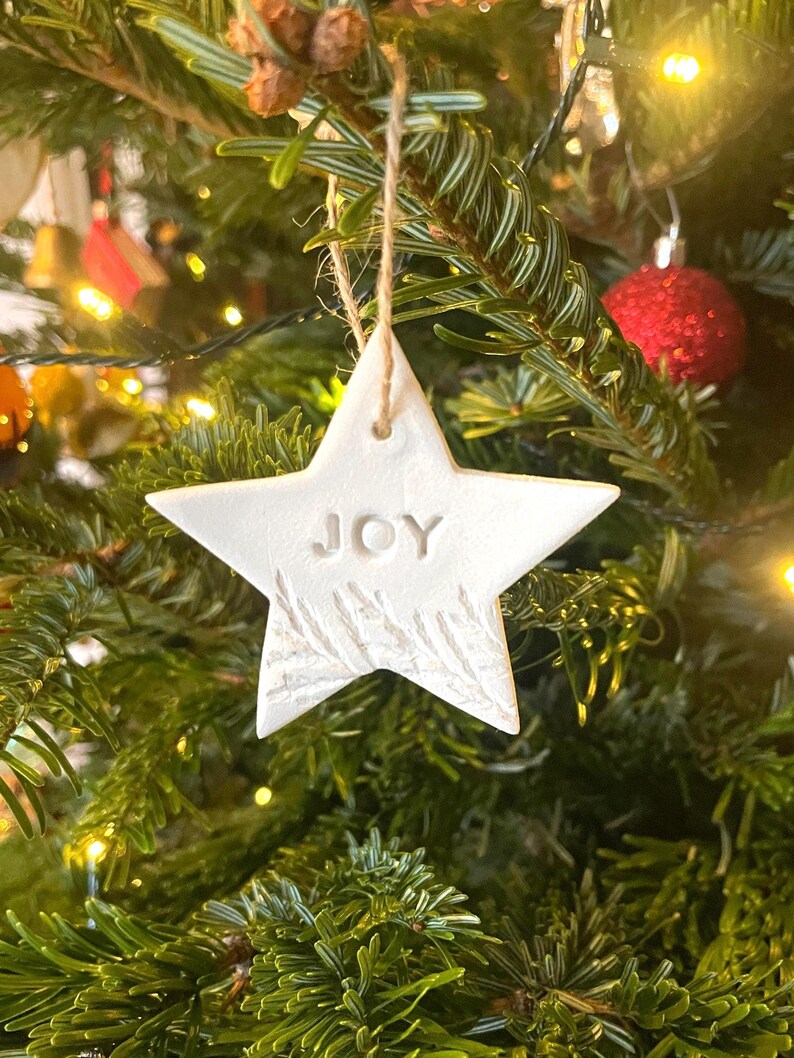 A white star-shaped Christmas ornament with the word "JOY" embossed on it. The ornament has a textured surface and a natural twine hanger. The star is hanging on a green Christmas tree with red baubles and lights.