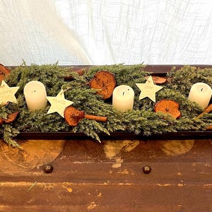 A rectangular wooden tray arrangement featuring greenery, dried fruit, cinnamon sticks, and four white pillar candles. White clay star decorations with the words "HOPE", "JOY", "PEACE", and "LOVE" are also included.