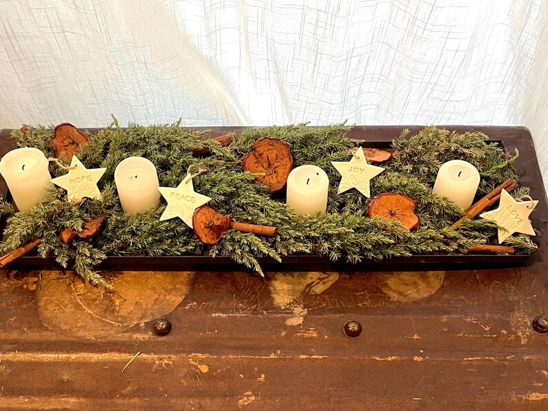A rectangular wooden tray arrangement featuring greenery, dried fruit, cinnamon sticks, and four white pillar candles. White clay star decorations with the words "HOPE", "JOY", "PEACE", and "LOVE" are also included.