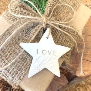 A wrapped gift with a burlap ribbon and a white star-shaped ornament. The ornament has the word "LOVE" engraved on it. The gift is decorated with a sprig of green foliage and sits on a wooden surface.