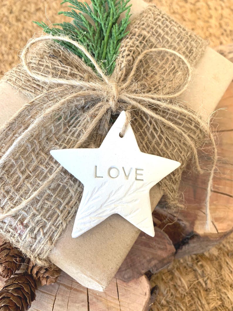 A wrapped gift with a burlap ribbon and a white star-shaped ornament. The ornament has the word "LOVE" engraved on it. The gift is decorated with a sprig of green foliage and sits on a wooden surface.