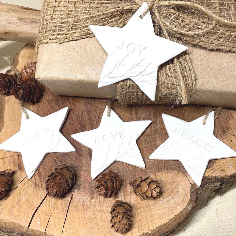 White star-shaped decorations with the words "JOY", "HOPE", "LOVE", and "PEACE" embossed. The ornaments feature a leaf design and are suspended by twine. They are arranged with pine cones and a wrapped gift on a wooden surface.