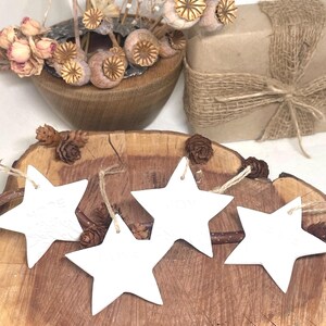 Four white star-shaped decorations with twine hangers are displayed on a rustic wooden surface. The stars are arranged in a row, with a natural wood background. Dried flowers and a wrapped gift are in the background.