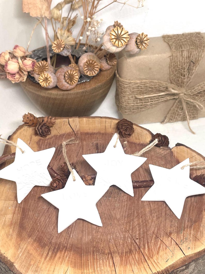 Four white star-shaped decorations with twine hangers are displayed on a rustic wooden surface. The stars are arranged in a row, with a natural wood background. Dried flowers and a wrapped gift are in the background.