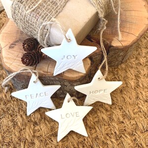 Four white star-shaped decorations, each with a word embossed: "JOY", "PEACE", "LOVE", and "HOPE". They have hanging strings. A wrapped gift with a burlap bow and pine cones are in the background.