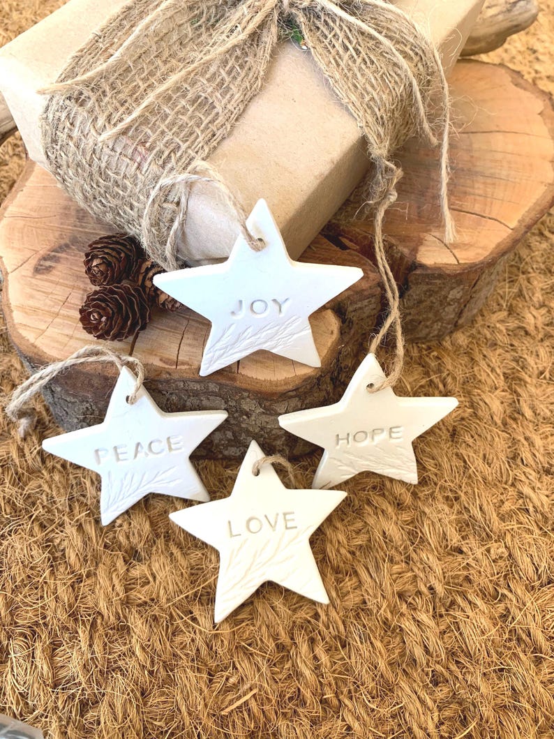 Four white star-shaped decorations, each with a word embossed: "JOY", "PEACE", "LOVE", and "HOPE". They have hanging strings. A wrapped gift with a burlap bow and pine cones are in the background.