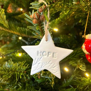 A white star-shaped Christmas decoration with the word "HOPE" embossed on it. The ornament hangs from a natural twine string against a backdrop of green pine needles and small lights. The star has a textured leaf design.
