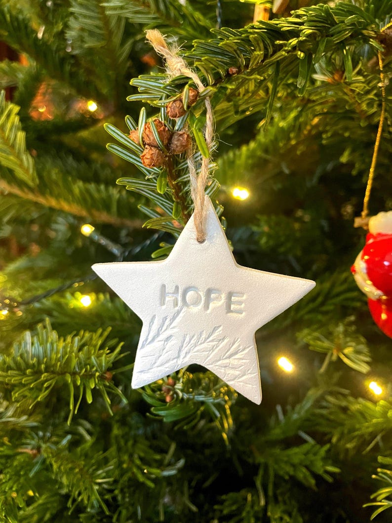 A white star-shaped Christmas decoration with the word "HOPE" embossed on it. The ornament hangs from a natural twine string against a backdrop of green pine needles and small lights. The star has a textured leaf design.