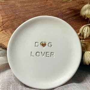 May include: A white ceramic dish with the words "DOG LOVER" embossed on the surface. A gold heart is between the words "DOG" and "LOVER". The dish is round and sits on a white cloth.