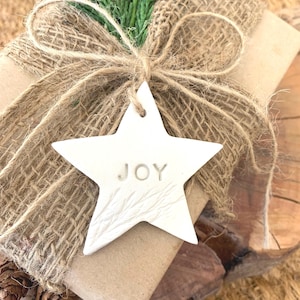 A wrapped gift with a white star-shaped tag that reads "JOY". The gift is wrapped in brown paper and burlap, tied with twine, and adorned with a sprig of green foliage. Pinecones are visible in the background.