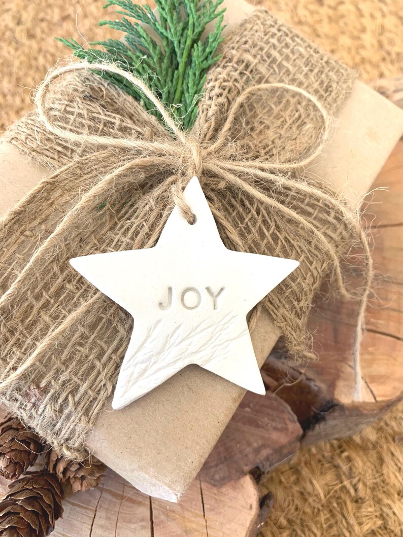A wrapped gift with a white star-shaped tag that reads "JOY". The gift is wrapped in brown paper and burlap, tied with twine, and adorned with a sprig of green foliage. Pinecones are visible in the background.