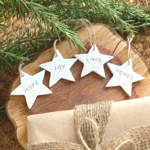 Four white star-shaped decorations, each inscribed with a word: "HOPE", "JOY", "LOVE", and "PEACE". They hang from twine and are arranged on a wooden surface. A wrapped gift with burlap ribbon and rosemary sprig are also present.