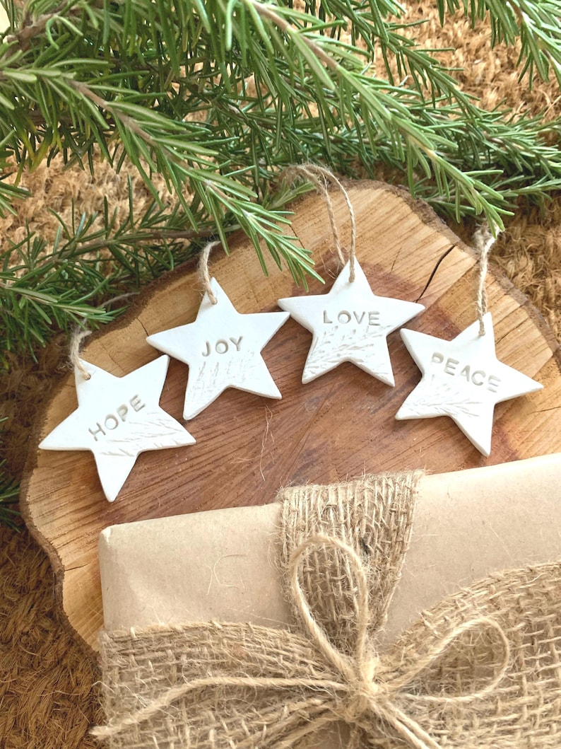 Four white star-shaped decorations, each inscribed with a word: "HOPE", "JOY", "LOVE", and "PEACE". They hang from twine and are arranged on a wooden surface. A wrapped gift with burlap ribbon and rosemary sprig are also present.