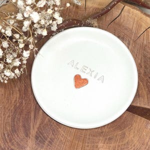 May include: A round, off-white ceramic trinket dish featuring the name ALEXIA embossed in capital letters above a small, textured copper-coloured heart shape centred on the smooth, flat surface of the circular tray.