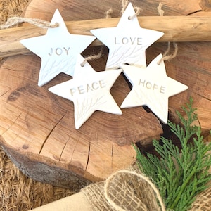 Four white star-shaped decorations, each inscribed with the words "JOY", "LOVE", "PEACE", and "HOPE". They are suspended by twine loops. The ornaments are displayed on a wooden surface, with a natural, rustic appearance, and a sprig of green foliage.