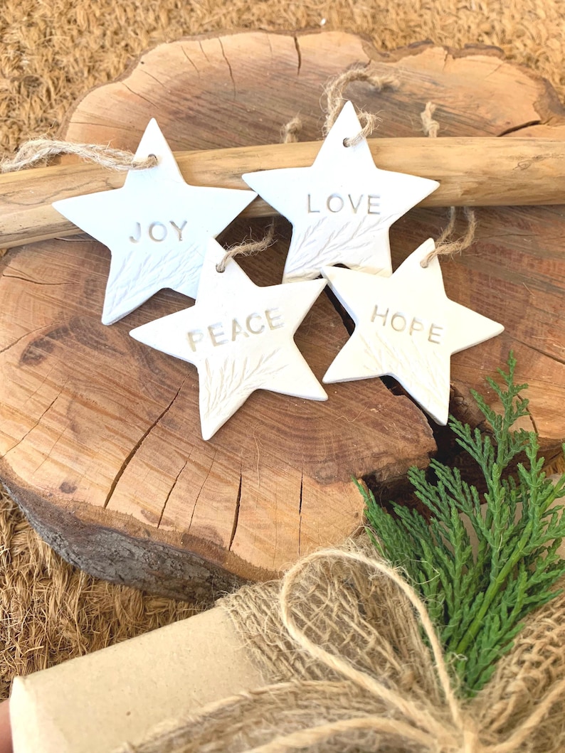 Four white star-shaped decorations, each inscribed with the words "JOY", "LOVE", "PEACE", and "HOPE". They are suspended by twine loops. The ornaments are displayed on a wooden surface, with a natural, rustic appearance, and a sprig of green foliage.