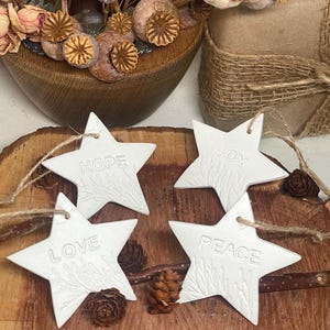 Four white star-shaped ornaments with the words "HOPE", "JOY", "LOVE", and "PEACE" embossed on them. Each star has a textured design and is strung with twine. The ornaments are displayed on a wooden surface.