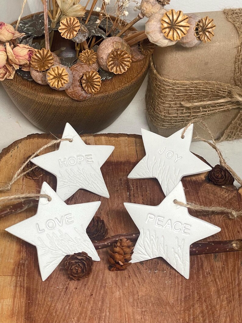 Four white star-shaped ornaments with the words "HOPE", "JOY", "LOVE", and "PEACE" embossed on them. Each star has a textured design and is strung with twine. The ornaments are displayed on a wooden surface.