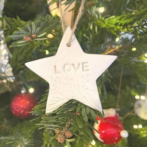 A white star-shaped Christmas decoration with the word "LOVE" embossed on it, suspended by a natural twine string. The ornament is set against a decorated Christmas tree with red and white baubles.