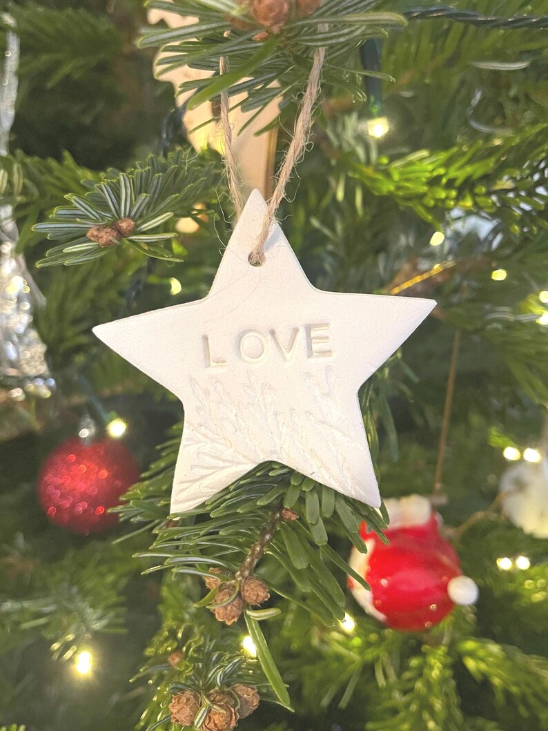 A white star-shaped Christmas decoration with the word "LOVE" embossed on it, suspended by a natural twine string. The ornament is set against a decorated Christmas tree with red and white baubles.