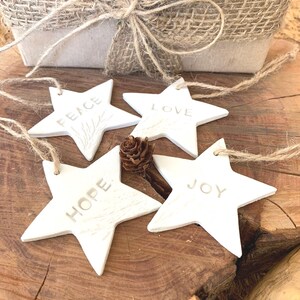 Four white star-shaped decorations, each with a word embossed: "PEACE", "LOVE", "HOPE", and "JOY". They are strung with twine and lie on a wooden surface. A small pine cone and a wrapped gift box are in the background.