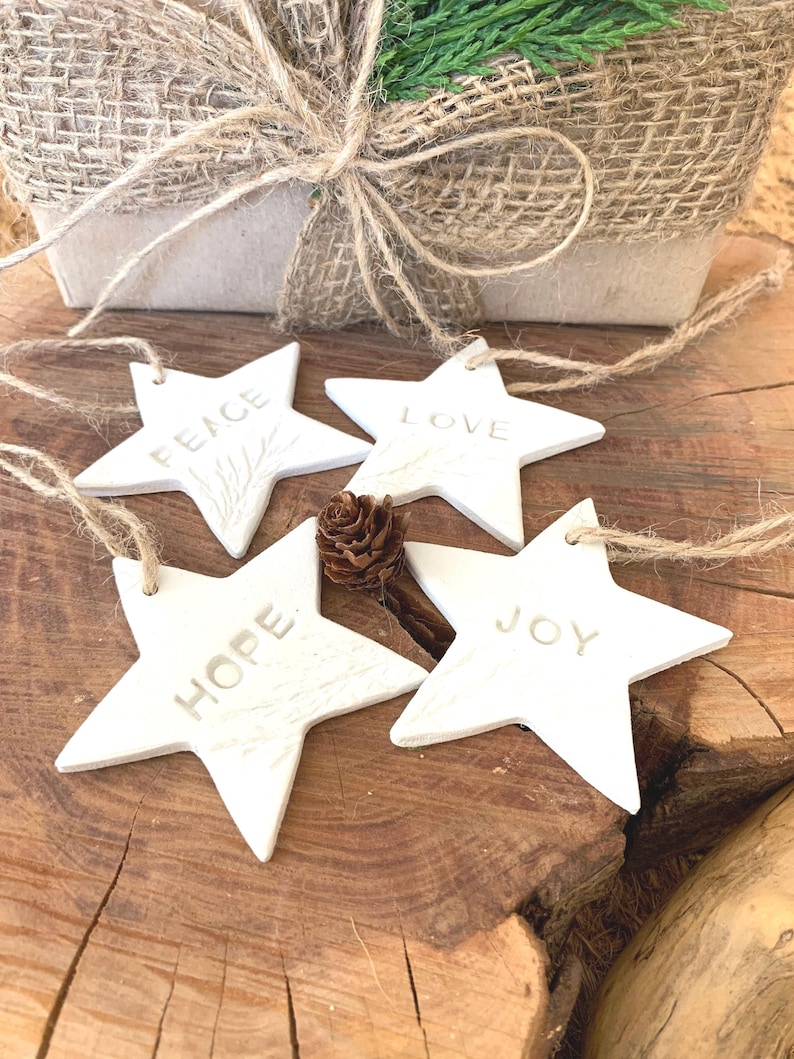 Four white star-shaped decorations, each with a word embossed: "PEACE", "LOVE", "HOPE", and "JOY". They are strung with twine and lie on a wooden surface. A small pine cone and a wrapped gift box are in the background.