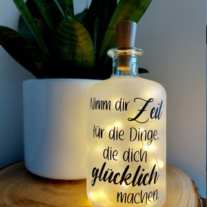 Könnte beinhalten: Eine gefrostete Glasflasche mit einem Korkverschluss und einer Lichterkette im Inneren. Die Flasche hat ein Zitat darauf gedruckt, das lautet: "Nimm dir Zeit für die Dinge, die dich glücklich machen."