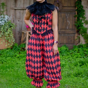 May include: A sleeveless jumpsuit featuring a red and black diamond harlequin pattern, a black ruffled collar, and a lace-up bodice with metal grommets. The garment has a fitted waist and loose, gathered pant legs that end in ruffled cuffs.