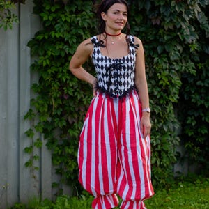 May include: A sleeveless corset top featuring a black and white diamond harlequin pattern with black ribbon shoulder ties and a front lace-up closure, paired with loose-fitting red and white vertical striped trousers with ruffled hem cuffs.