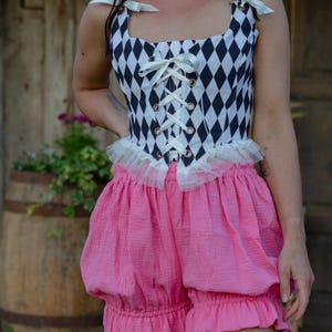 May include: A black and white diamond-patterned corset top featuring a front lace-up closure with white ribbons, shoulder ties, and a white tulle ruffle at the waist, paired with bright pink, high-waisted, loose-fitting bloomers with ruffled hems.