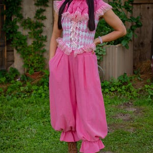 May include: A pink and white diamond-patterned corset top with a ruffled collar and lace-up front, paired with loose-fitting pink trousers featuring ruffled hems and side pockets, worn with brown lace-up boots.