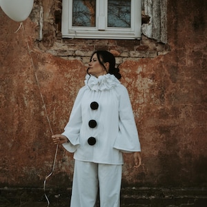 May include: A white Pierrot costume with a ruffled collar and three large black buttons. The outfit includes wide-leg pants and flared sleeves. A white balloon is held by a string. The background is a weathered brick wall.