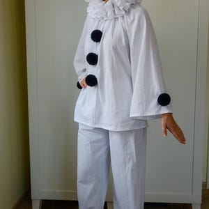 May include: White clown costume featuring a ruffled collar, large black pom-poms, and wide-leg pants. The costume includes a long-sleeved top and is suitable for theatrical performances or costume parties.