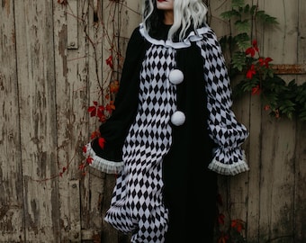 May include: A long-sleeved clown costume featuring a black and white diamond-patterned bodice and solid black sleeves. The garment includes a ruffled collar, two large white pom-poms on the front, and white ruffled cuffs at the wrists.