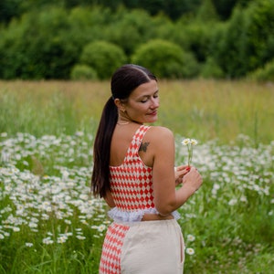 May include: A woman wearing a red and white checkered crop top with a white ruffle trim and matching pants. She is holding a small bouquet of white daisies in her right hand. The woman is standing in a field of daisies.
