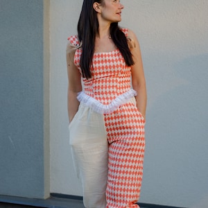 May include: A woman wearing a red and white checkered jumpsuit with a white ruffle trim. The jumpsuit has a white fabric panel on the left leg. She is standing in front of a light gray wall.
