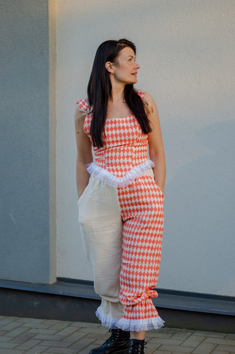 May include: A woman wearing a red and white checkered jumpsuit with a white ruffle trim. The jumpsuit has a white fabric panel on the left leg. She is standing in front of a light gray wall.