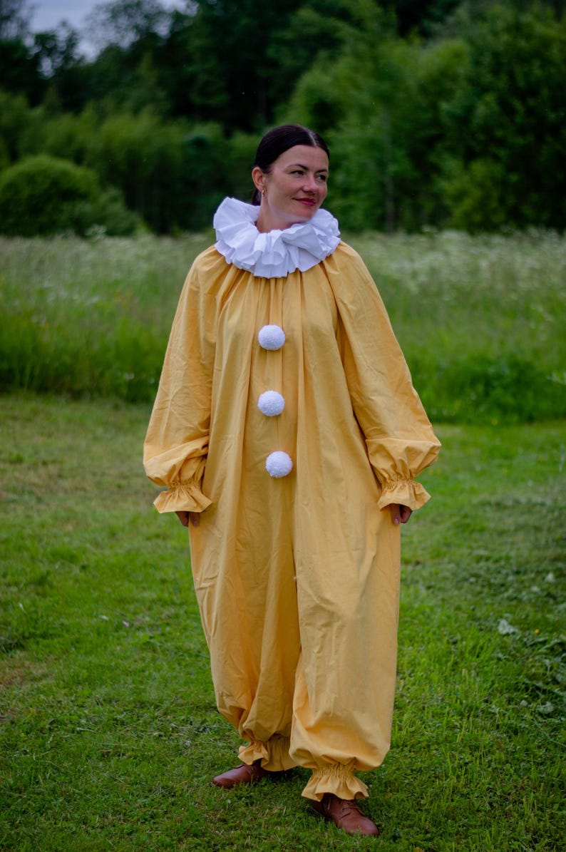 May include: A yellow clown costume with a white ruffled collar and three white pom-poms down the front. The long-sleeved jumpsuit has ruffled cuffs and is worn with brown shoes. The person is standing in a grassy field.