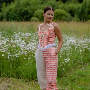 May include: A woman wearing a red and white checkered top with a white ruffle and matching pants. The pants are split, with one leg being white and the other red and white checkered. She is standing in a field of white flowers.
