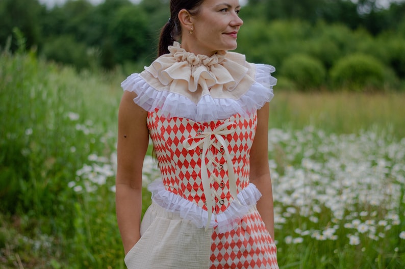 May include: A woman wearing a white and red checkered corset with a white lace trim and a white ruffled collar. The corset is tied with a white ribbon. She is standing in a field of white flowers.